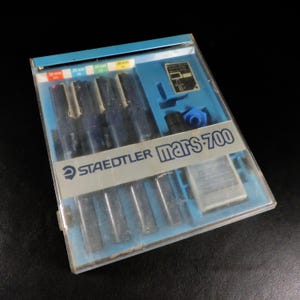 May include: A Staedtler Mars 700 technical pen set in a clear plastic case. The set includes several black pens with varying tip sizes, indicated by colored markings. The case has a blue interior and a label with the Staedtler logo.
