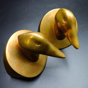 May include: A pair of gold-toned metal bookends shaped like duck heads. The duck heads are facing forward and have a smooth, rounded surface.