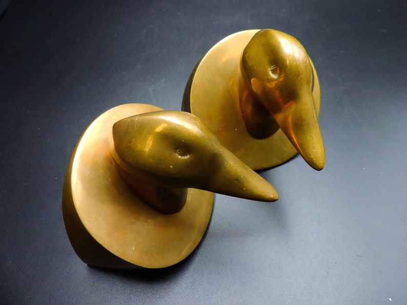 May include: A pair of gold-toned metal bookends shaped like duck heads. The duck heads are facing forward and have a smooth, rounded surface.