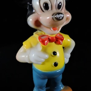 May include: A vintage Mickey Mouse figurine. The figure is wearing a yellow shirt with black buttons, blue trousers, a red bow tie, and orange shoes. The figurine has black ears and a white face with rosy cheeks. It stands on a blue base.