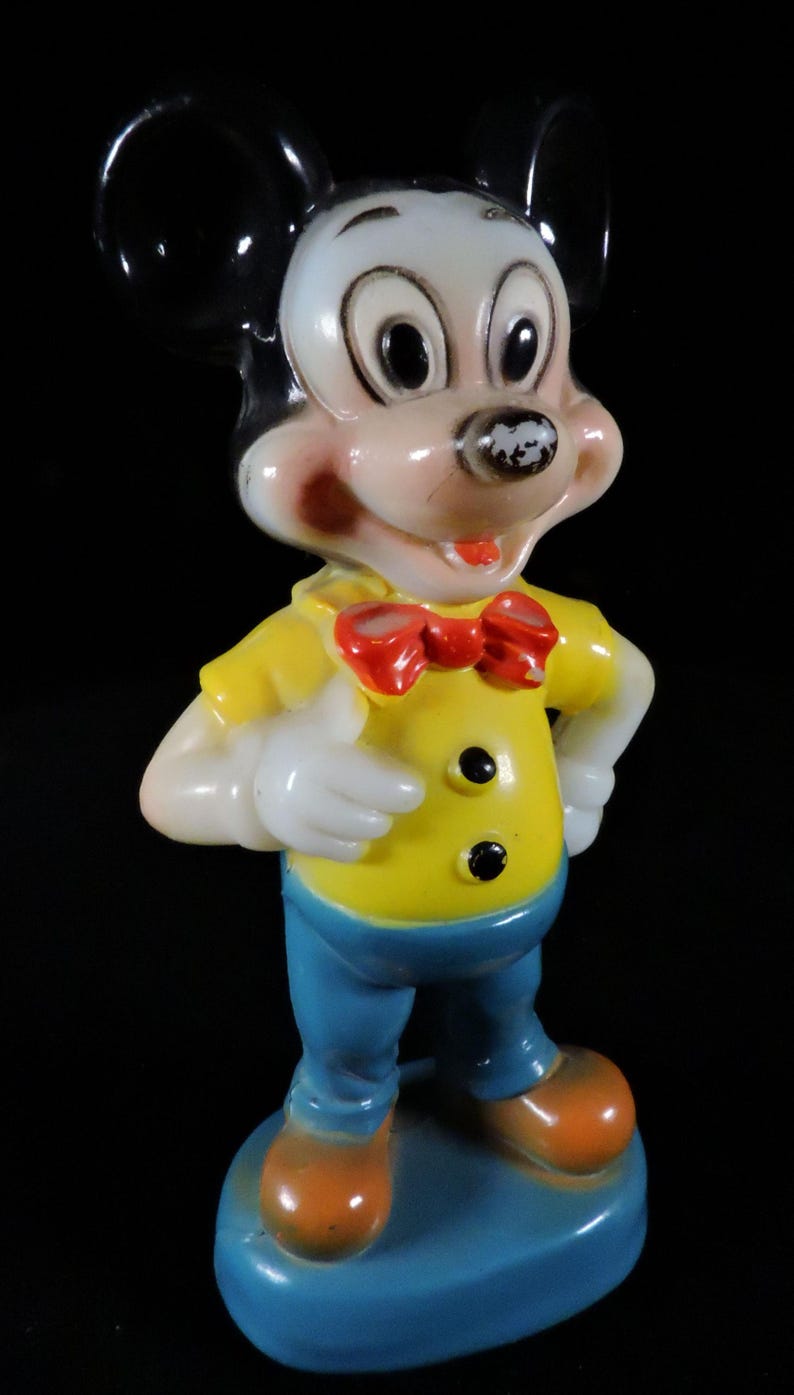 May include: A vintage Mickey Mouse figurine. The figure is wearing a yellow shirt with black buttons, blue trousers, a red bow tie, and orange shoes. The figurine has black ears and a white face with rosy cheeks. It stands on a blue base.