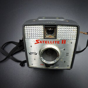 Könnte beinhalten: Eine schwarze und silberne Vintage-Kamera mit einem schwarzen Band. Die Kamera hat ein Objektiv mit einem silbernen Rand und den Text "SATELLITE II" in roten Buchstaben. Die Kamera hat auch den Text "Imperial" und "127 4x4" in schwarzen Buchstaben.