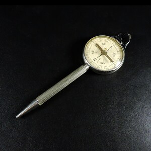 May include: A silver-toned metal pen with a circular compass integrated into the top. The compass features a cream-colored face with black cardinal directions, degree markings, and a central needle. The pen body has a textured, ribbed grip.