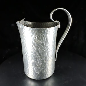 May include: A silver pitcher with a hammered finish and floral design. The pitcher has a curved handle and a wide mouth.