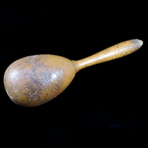 May include: A wooden egg-shaped maraca with a long, brown handle. The maraca has a rough, textured surface.
