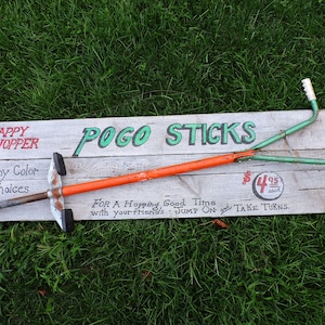 May include: A vintage Pogo Stick with a green and orange frame, resting on a wooden sign that reads "POGO STICKS" and "HAPPY HOPPER". The sign also includes the price "$4.95 each" and the text "FOR A Hopping Good Time with your friends - JUMP ON & TAKE TURNS."