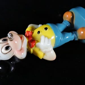 May include: A vintage Mickey Mouse figurine in a lying down pose. The figure has a white face, black ears, and a yellow body with blue trousers. It has orange feet and a red bow tie. The figurine is on a black surface.