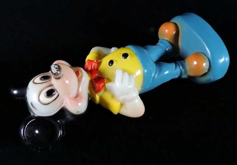 May include: A vintage Mickey Mouse figurine in a lying down pose. The figure has a white face, black ears, and a yellow body with blue trousers. It has orange feet and a red bow tie. The figurine is on a black surface.