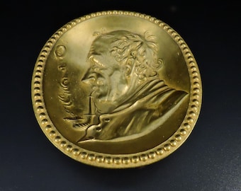 Op de afbeelding: Een gouden metalen medaillon met een reliëfprofiel van een mannengezicht. Het medaillon heeft een parelrand en de inscriptie "O.C.A." aan de linkerkant.