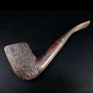 Peut inclure: Une pipe à tabac vintage avec un foyer brun foncé texturé et un tuyau incurvé. La tige est brun clair avec une bague argentée près du foyer. La pipe est conçue pour fumer du tabac.