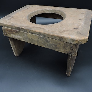 May include: A rustic wooden stool with a circular cutout in the top. The stool is made of weathered wood and has a simple design.