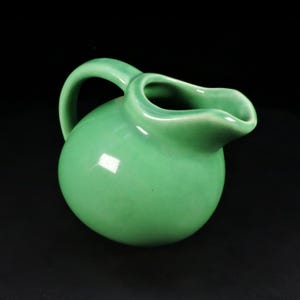 May include: A small, round ceramic pitcher with a smooth, glossy light green glaze. The vessel features a curved handle on the side and a wide, integrated pouring spout at the top, showcasing a uniform color and a reflective, polished surface finish.