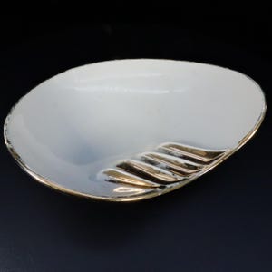 May include: White ceramic ashtray with a gold-colored, ribbed design. The ashtray is oval-shaped and has a slight curve.