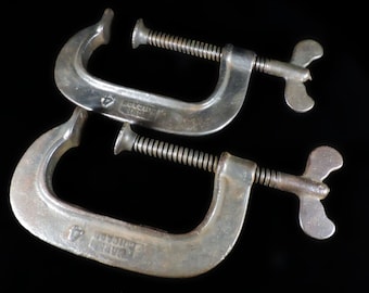 May include: Two vintage C-clamps made of silver-coloured metal. Each clamp has a threaded screw and a butterfly-shaped handle for tightening. The clamps are marked with the text "WARREN CHICAGO 4".