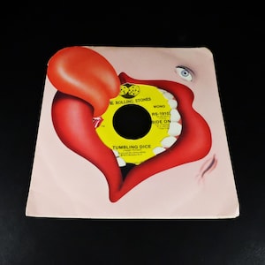 May include: A vintage vinyl record sleeve featuring the Rolling Stones' "Tumbling Dice." The artwork showcases a red tongue and lips design with a yellow record label visible in the center. The sleeve has a light pink background and the text "Tumbling Dice" is visible.