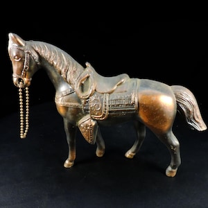 May include: A bronze-coloured horse figurine with a saddle and bridle. The horse has a detailed mane and tail, and a beaded chain hangs from its mouth. The figurine is set against a black background.
