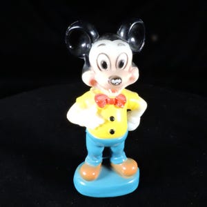May include: A vintage Mickey Mouse figurine. The figure is wearing a yellow shirt with black buttons, blue trousers, and orange shoes. A red bow tie and black ears complete the look. The figurine stands on a blue base.