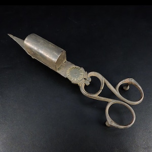 May include: Antique silver-toned scissors with a cylindrical blade housing and ornate handle. The handle features two finger loops and a decorative floral element. The scissors are set against a dark background.