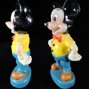 May include: Vintage Mickey Mouse figurine. The figure is wearing a yellow shirt, blue trousers, and orange shoes. It has a red bow tie and black ears. The figurine is standing on a blue base. The figure is made of plastic.