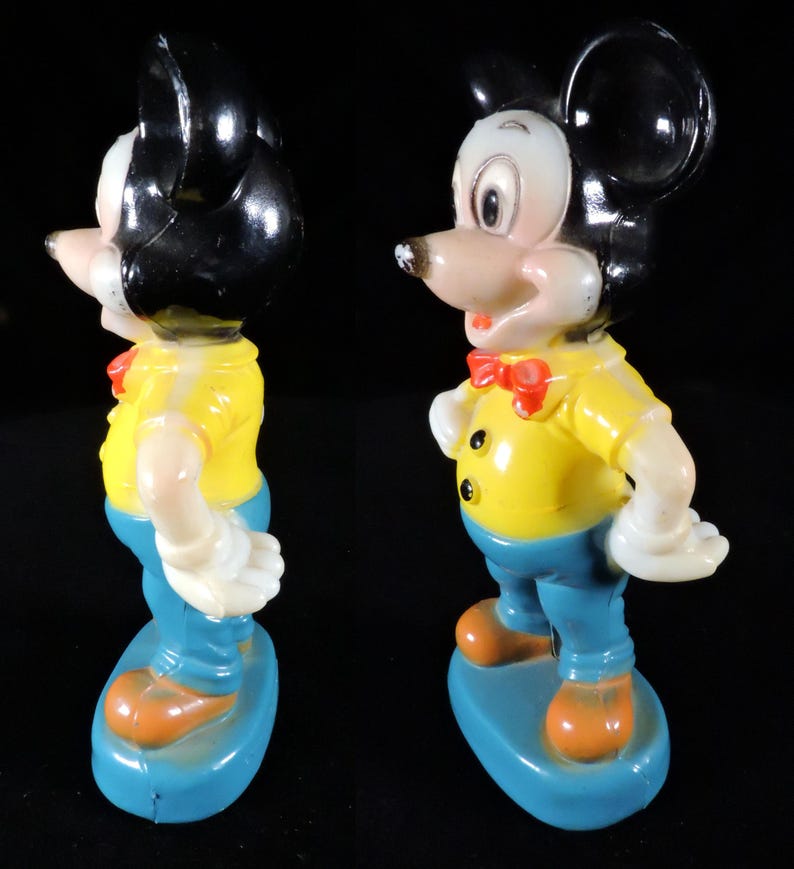 May include: Vintage Mickey Mouse figurine. The figure is wearing a yellow shirt, blue trousers, and orange shoes. It has a red bow tie and black ears. The figurine is standing on a blue base. The figure is made of plastic.