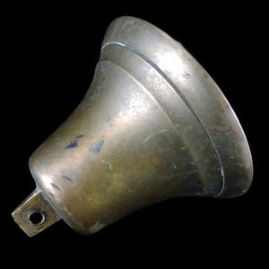 May include: A vintage brass bell with a flared shape and a small rectangular mounting bracket. The bell has a tarnished, aged appearance with visible wear and tear, and a dark background.