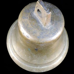 May include: A bronze-colored bell with a loop at the top for hanging. The bell has a rounded top and a flared base. The metal has a weathered appearance, with variations in color and texture. The background is black.