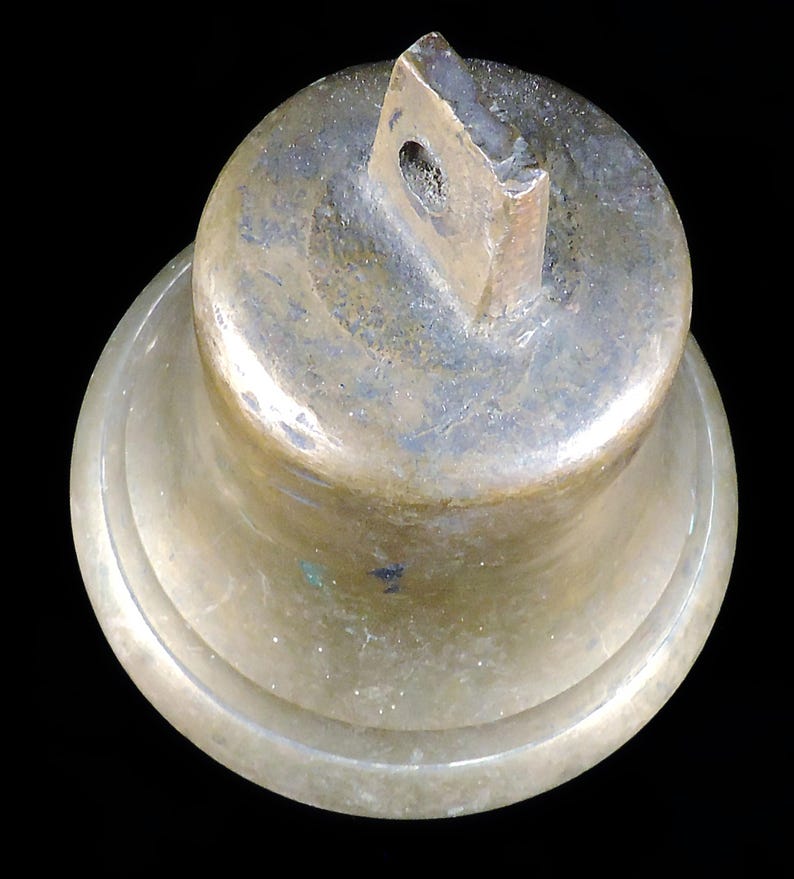 May include: A bronze-colored bell with a loop at the top for hanging. The bell has a rounded top and a flared base. The metal has a weathered appearance, with variations in color and texture. The background is black.