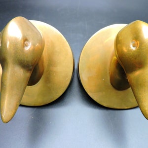 May include: A pair of gold-toned metal bookends shaped like duck heads. The bookends are on a dark background.