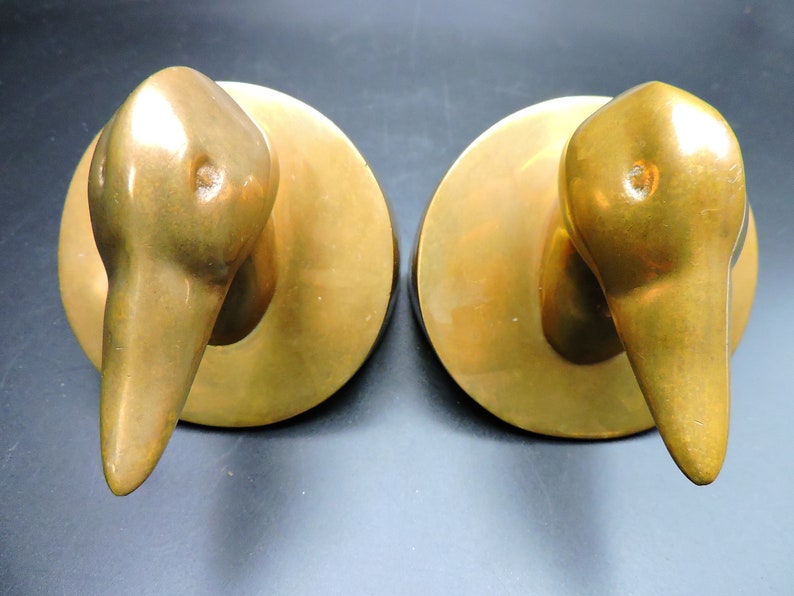 May include: A pair of gold-toned metal bookends shaped like duck heads. The bookends are on a dark background.