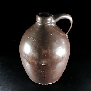 May include: A brown ceramic jug with a handle. The jug has a rounded body and a narrow neck. The surface is glossy and reflects light. The jug is likely used for storing liquids.
