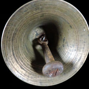 May include: A close-up view of a brass bell with a dark, aged patina. The bell's interior shows concentric circular ridges. A rusty, metal clapper hangs inside, with a rounded, knob-like end.