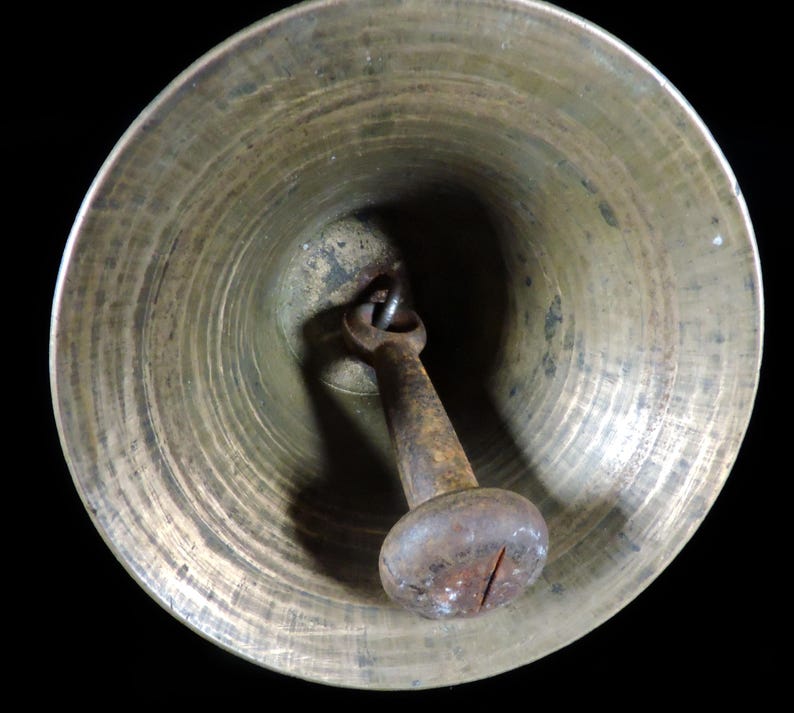 May include: A close-up view of a brass bell with a dark, aged patina. The bell's interior shows concentric circular ridges. A rusty, metal clapper hangs inside, with a rounded, knob-like end.
