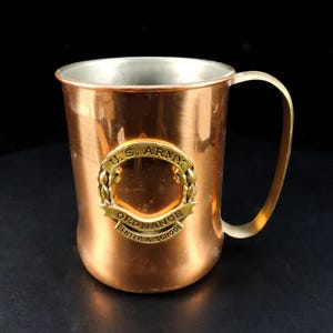 May include: A copper-colored mug with a gold-colored handle and a silver-colored interior. The mug features a gold-colored emblem with the text "U.S. ARMY ORDNANCE CENTER & SCHOOL".