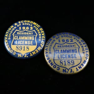 May include: Two round, blue and gold, metal buttons with the text "Division of Shell Fisheries Conservation & Economic Development" and "Resident Clamming License" with the year 1963 and 1962, and license numbers 8918 and 8189.