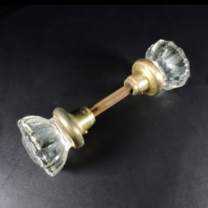 May include: A clear glass door knob with a faceted design and brass hardware. The knob is connected by a threaded rod. The hardware has a vintage appearance. The door knob is set against a black background.