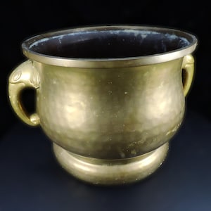 May include: A large, gold-colored metal planter with two elephant-shaped handles. The planter has a rounded bottom and a wide, open top.