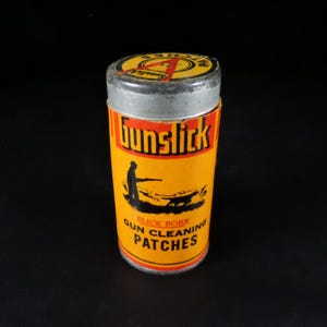 May include: A cylindrical container of "Gunslick" gun cleaning patches. The container is yellow with red and black text and graphics, including a silhouette of a person aiming a gun. The lid is silver.