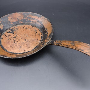 May include: A vintage copper frying pan with a black patina and a long handle. The pan has a hammered texture and shows signs of wear.