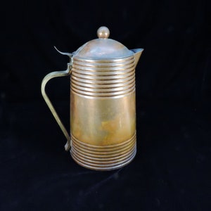 May include: A copper pitcher with a lid and a handle. The pitcher has a hammered finish and is decorated with horizontal lines.