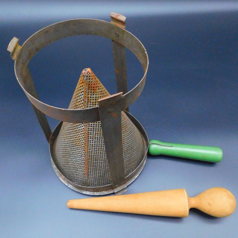 May include: A vintage metal food mill with a green handle and a wooden pestle. The mill is made of metal and has a conical shape with a mesh screen. The pestle is made of wood and has a rounded end.