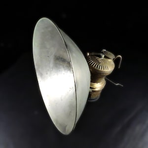 May include: Antique brass and silver-toned miner's lamp with a large, curved reflector. The lamp features a small, detailed oil reservoir and a curved handle. The reflector is oval-shaped and angled to direct light.