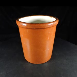 May include: A cylindrical ceramic crock featuring a smooth, glossy brown exterior glaze and a contrasting cream-coloured interior. The vessel has a thick, rounded rim at the top and tapers slightly towards the base, presenting a simple, functional design.