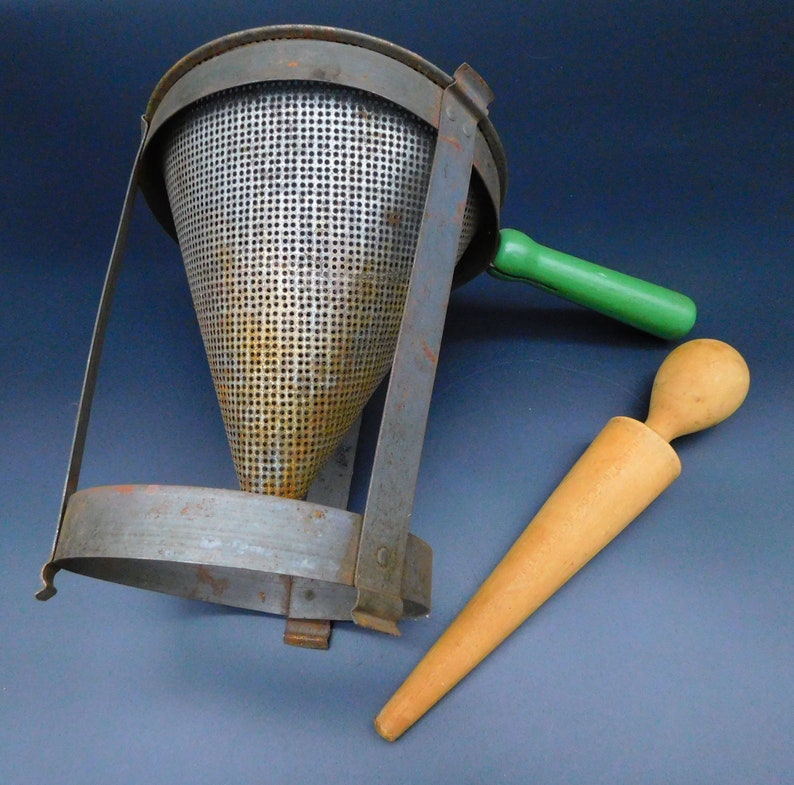 May include: A vintage metal food mill with a green handle and a wooden potato ricer. The food mill has a conical shape with a mesh screen and a metal frame. The ricer is long and slender with a rounded tip.