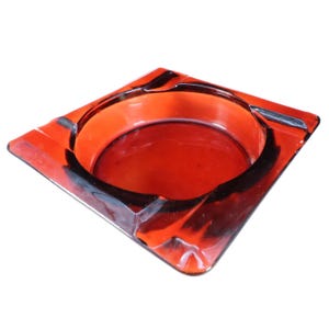 May include: A square-shaped ashtray crafted from translucent red glass. The design features a circular central basin surrounded by a flat, wide rim with four distinct notches positioned at each corner for resting items, showcasing a smooth finish.