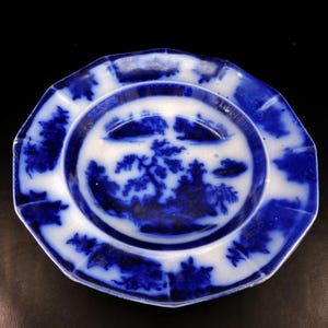May include: A blue and white antique plate with a landscape design. The plate has a scalloped edge and a central image of trees and a building. The plate's design is repeated around the rim. The plate is likely made of ceramic.