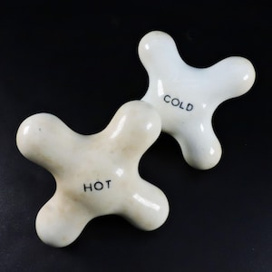 Peut inclure: Deux poignées de robinet en porcelaine blanc cassé, chacune avec un design à trois lobes. L'une est marquée "HOT" en noir, l'autre "COLD". Les poignées sont posées sur un fond noir.