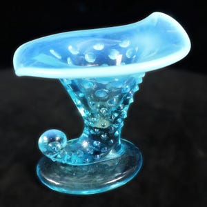 May include: A light blue, hobnail-style glass vase with a flared rim and a rounded base. The vase has a unique, curved shape with a small, spherical accent. The glass has a textured surface with small, raised dots.