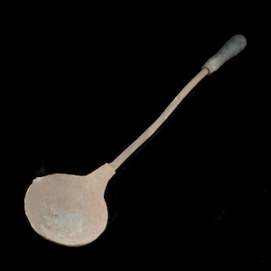 May include: A vintage metal ladle with a long, curved handle and a rounded bowl. The handle has a dark, textured grip. The ladle is made of a light-colored metal, possibly copper or brass, and shows signs of age and use.