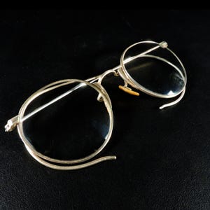 May include: Vintage round eyeglasses with a silver-colored metal frame. The glasses have clear lenses and small, curved temples. The nose pads are a light brown color. The glasses are set against a black background.