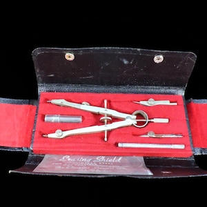 May include: An open black leather case with a red velvet interior, containing a set of drafting tools. The set includes a large compass, smaller dividers, a pencil holder, and a ruler. The tools are silver-colored.